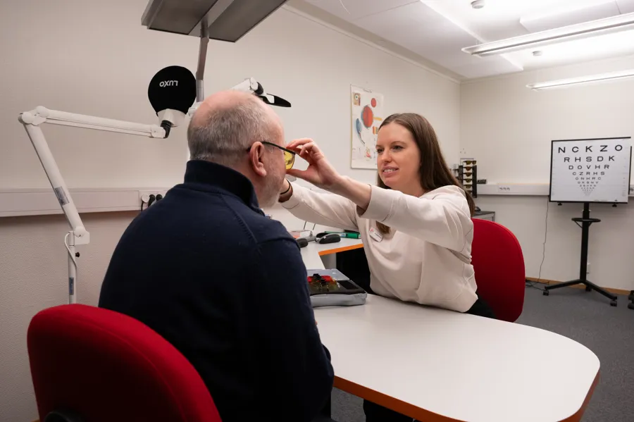 Mann sittende med svart genser med briller på seg og en kvinne med hvit trøye på andre siden av bordet holder på brillene.