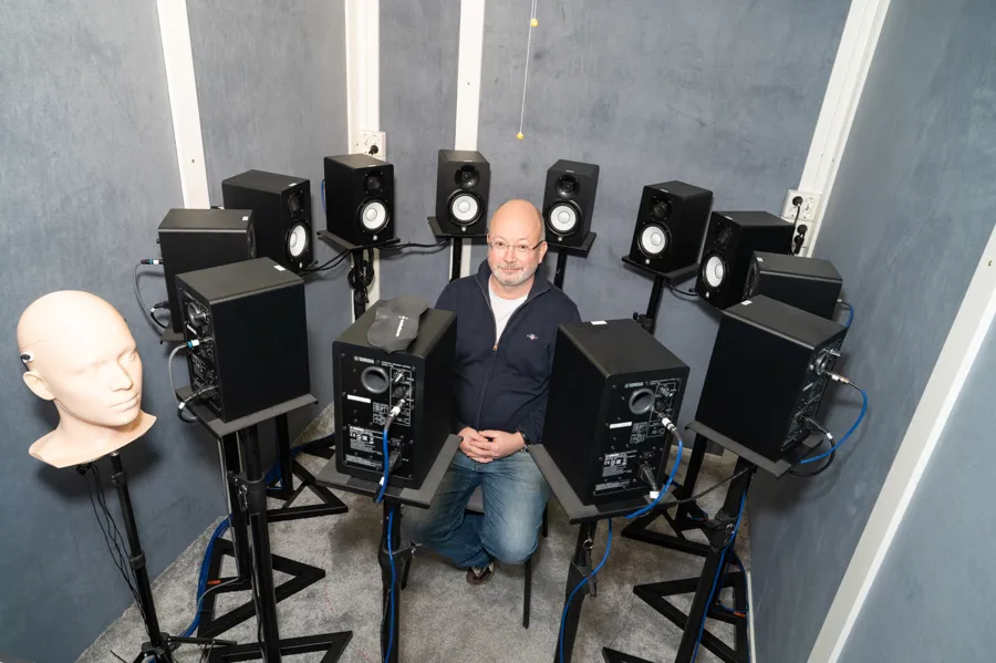 Mann med briller og svart trøye sittende med 12 høyttalere plassert rundt personen i hodehøyde.