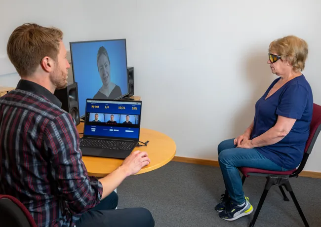 En mann sitter ved en PC og en kvinne med briller sitter ved siden av, hun ser på en TV-skjerm.