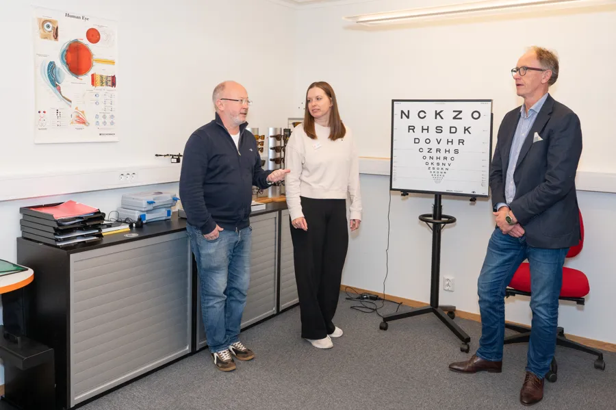 Tre personer stående ved siden av hverandre. På veggen er det plakat og en skjerm på hjul står på gulvet.