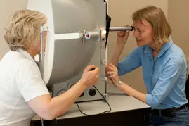 To kvinner der kvinnen til venstre har ansiktet inne i en maskin og kvinnen til høyre ser inn i maskinen og holder en hånd på et dreiehjul.