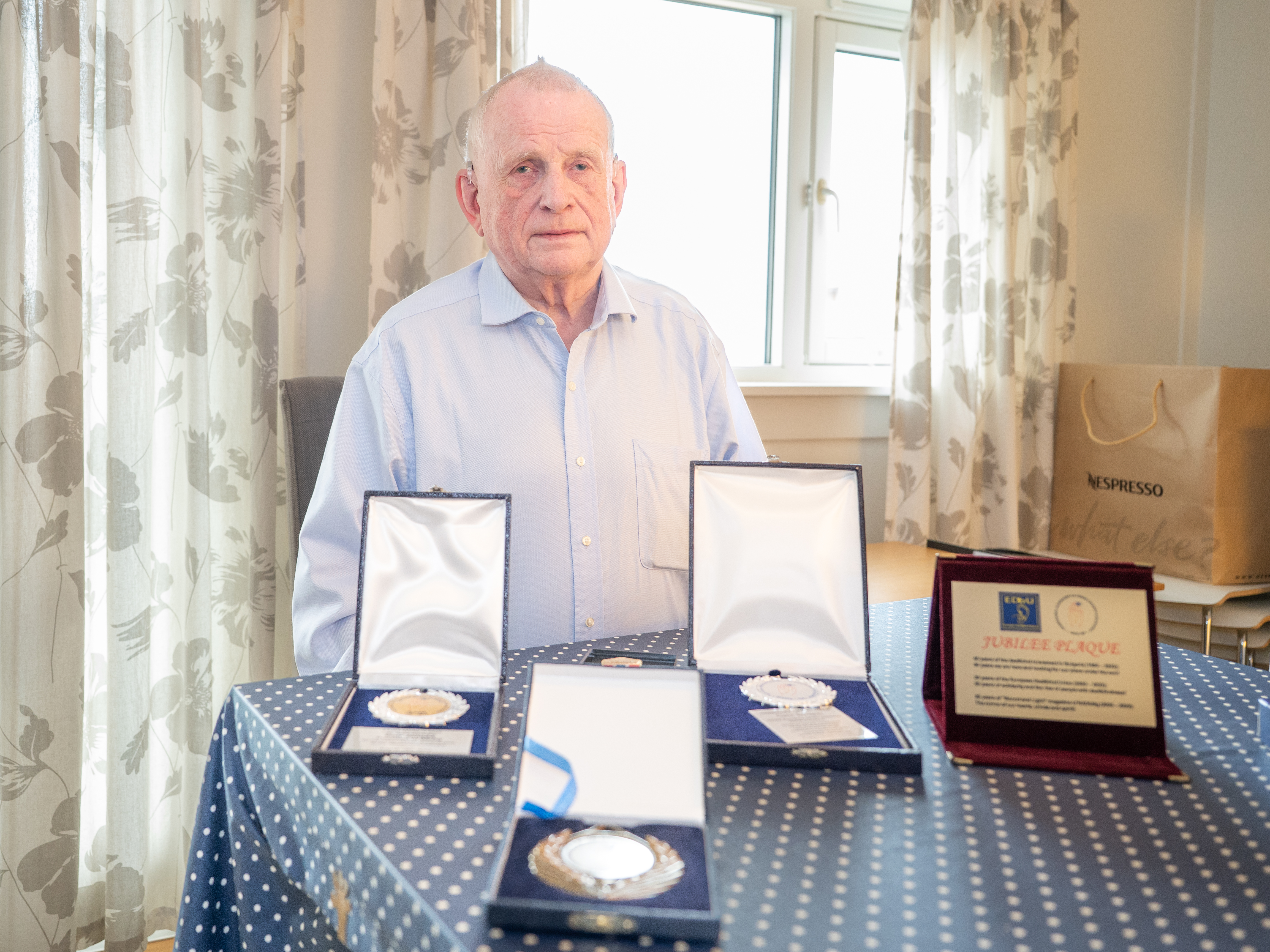 Eldre mann med blå skjorte sittende og på bordet foran ham er det plassert fire medaljer og et diplom.